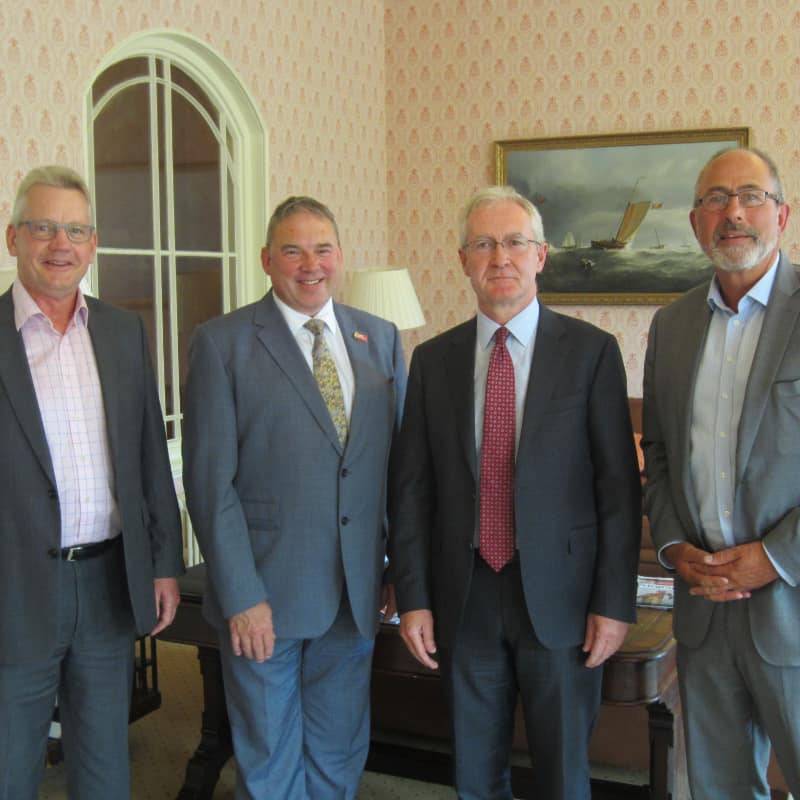 Four men in business attire standing in a room with decorative wallpaper and a painting in the background.