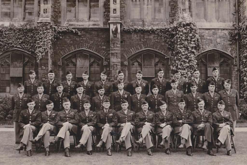 A group of uniformed military personnel posing for a formal black and white photograph in front of a historic building with ivy-covered walls.