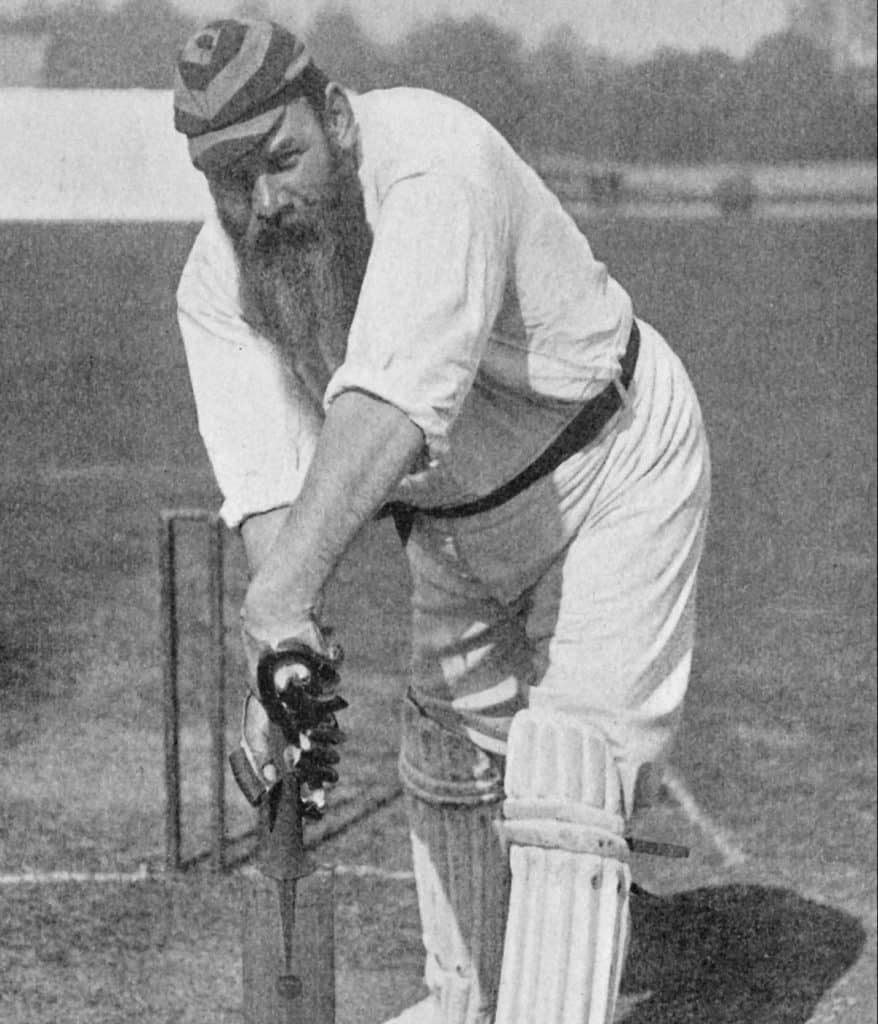 A vintage black-and-white photo of a bearded cricketer in traditional attire, preparing to bat.
