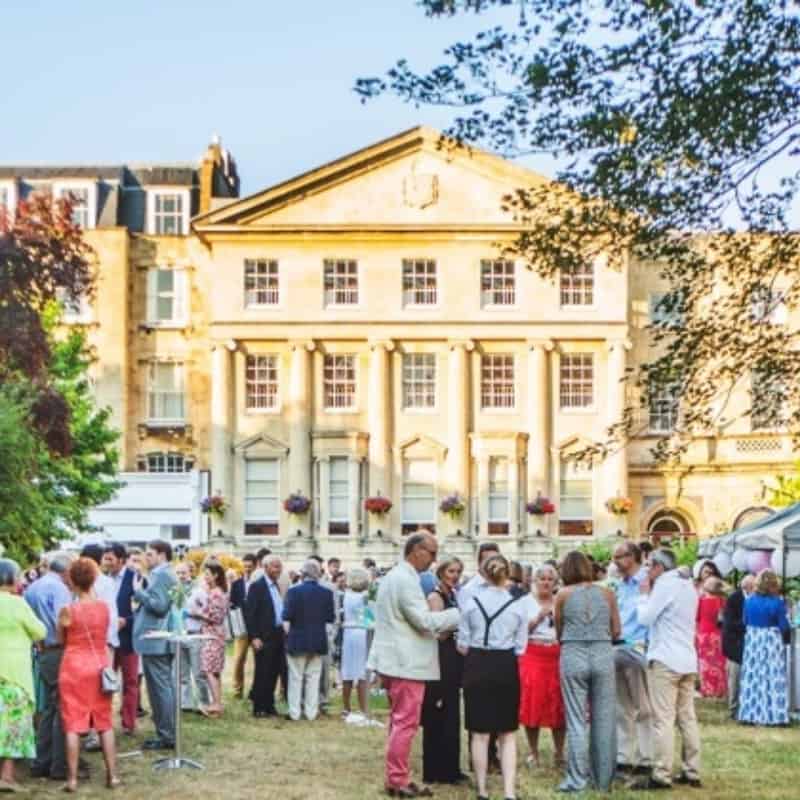 A social gathering outdoors with people conversing and enjoying a sunny day in front of an elegant building.