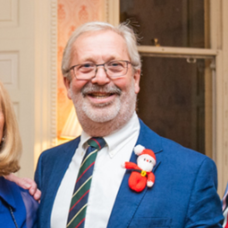 A smiling older man with gray hair, glasses, and a beard wears a blue suit, striped tie, and a festive Santa Claus pin on his jacket, standing indoors near a window and lamp.