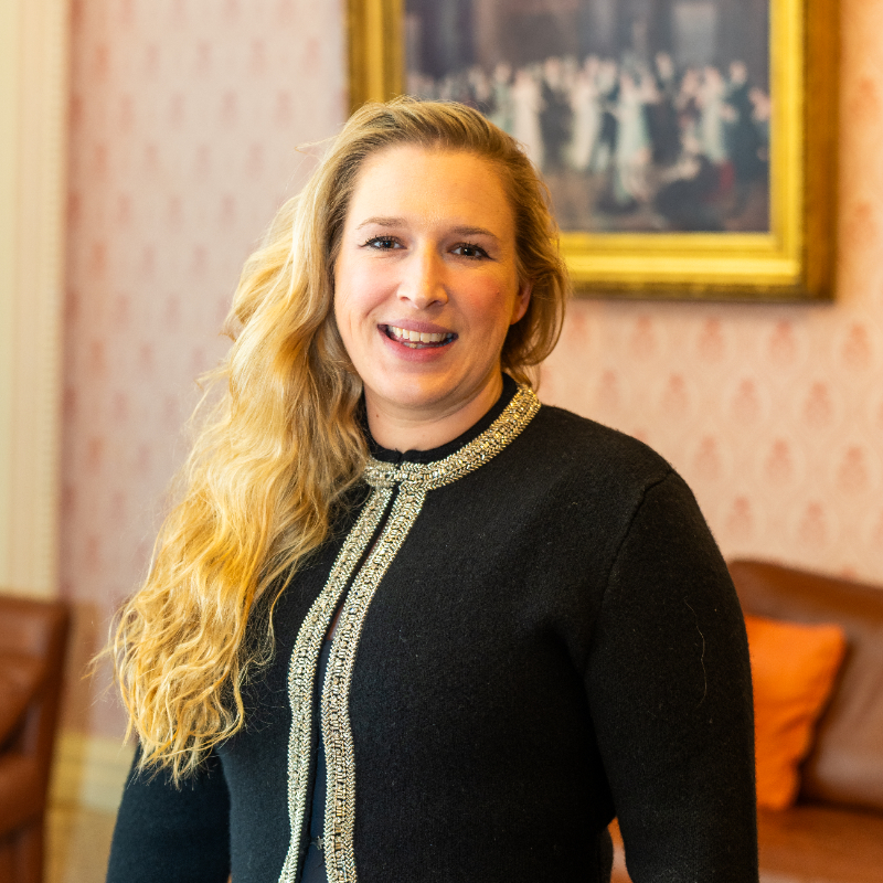 A woman with long blonde hair wearing a black jacket with a decorative trim stands indoors, smiling. Behind her is a patterned wall, a brown couch, and a framed painting.
