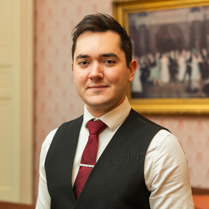 A young man with short dark hair wearing a white shirt, red tie, and black vest stands indoors, smiling slightly. A framed painting and patterned wallpaper are visible in the background.