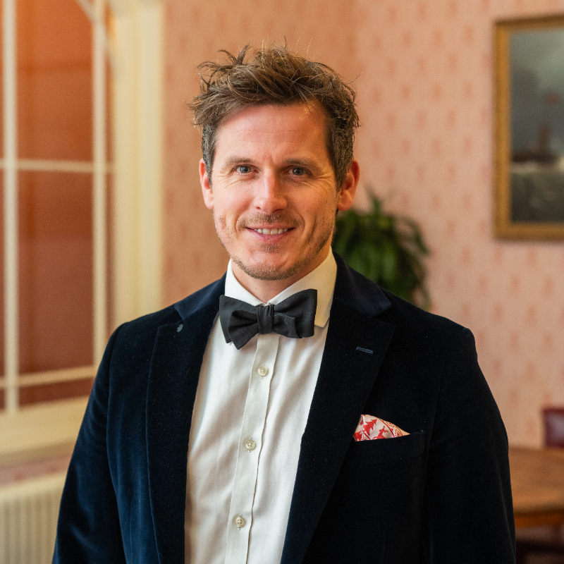 A man with short, tousled hair wearing a black velvet tuxedo jacket, white shirt, and black bow tie stands indoors, smiling. The background features a large window, a plant, and framed artwork on a patterned wall.