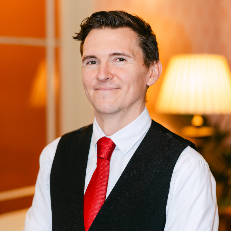 A man with short brown hair, wearing a white shirt, black waistcoat, and red tie, smiles at the camera in a warmly lit room with a lamp in the background.