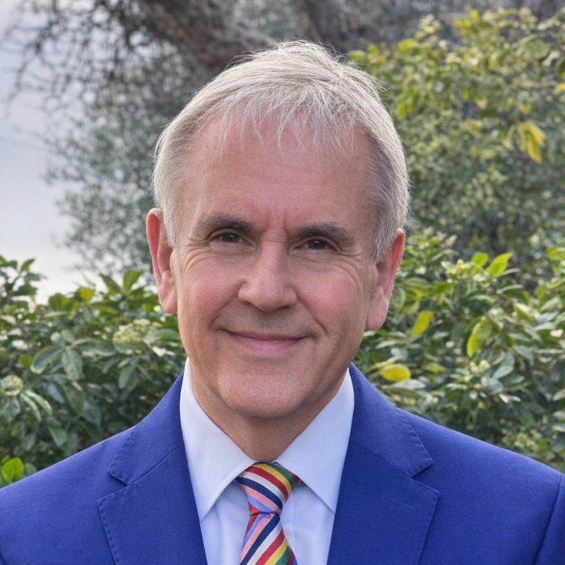 A smiling man with short grey hair wears a blue suit, white shirt, and colourful striped tie, standing outdoors in front of green foliage and trees.