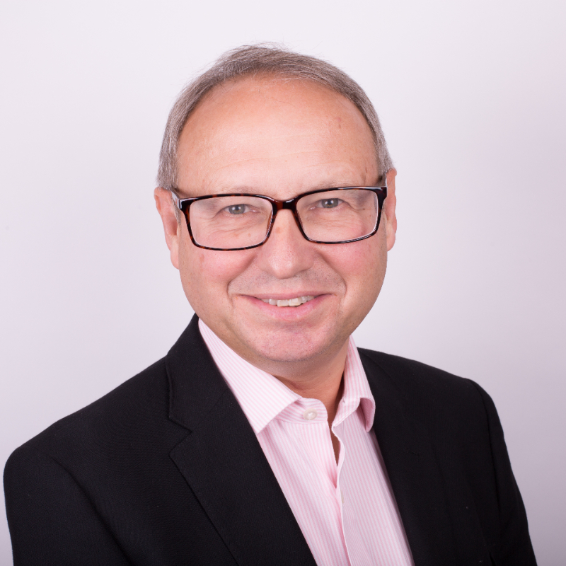 A middle-aged man wearing glasses, a black blazer, and a light pink collared shirt smiles at the camera against a plain light background.