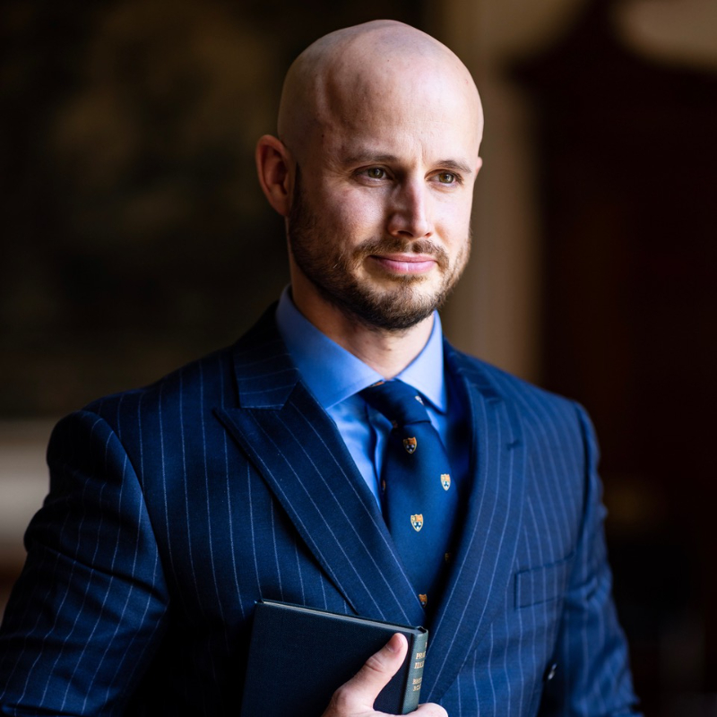 A bald man with a trimmed beard wearing a blue pinstripe suit, blue shirt, and patterned tie, holds a closed book and looks slightly to the side, standing in a softly lit indoor setting.