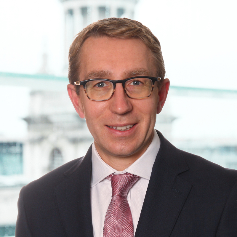 A man wearing glasses, a dark suit, white shirt, and light pink tie is smiling at the camera. The background is bright with an out-of-focus building visible through a window.