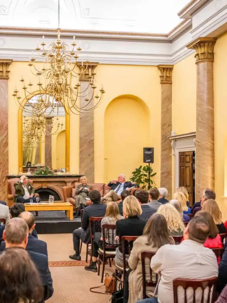 A panel discussion with three speakers seated on a stage, viewed from the back of an audience in a large, high-ceilinged room with yellow walls and a chandelier.